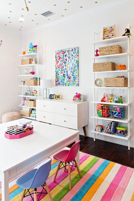 Custom playroom design with white wall shelving, woven baskets, pink and blue kids chairs, rainbow striped rug and gold star ceiling